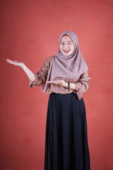 Beautiful Asian woman in brown shirt and hijab smiling cheerful presenting and pointing with palm of hand on brown background