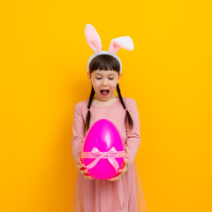 Cute baby little girl with bunny ears holding a big Easter egg on a colored yellow background