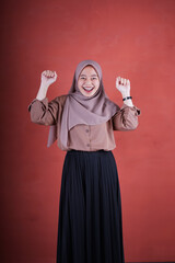 Beautiful Asian woman in brown shirt and hijab smiling cheerful very excited raising both hands on brown background