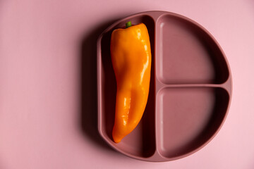 Top view of baby dark pink silicone plate divided on three zones with orange pepper on the pink background. First baby organic healthy self-feeding. 