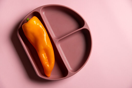 Top View Of Baby Dark Pink Silicone Plate Divided On Three Zones With Orange Pepper On The Pink Background. First Baby Organic Healthy Self-feeding. 