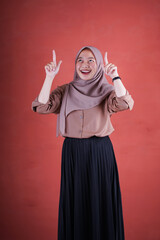Fototapeta na wymiar eautiful Asian woman in brown shirt and hijab smiling cheerful pointing up on brown background