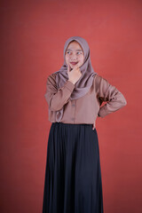 Fototapeta premium Beautiful Asian woman in brown shirt and hijab showing thinking expression with hand on chin on brown background