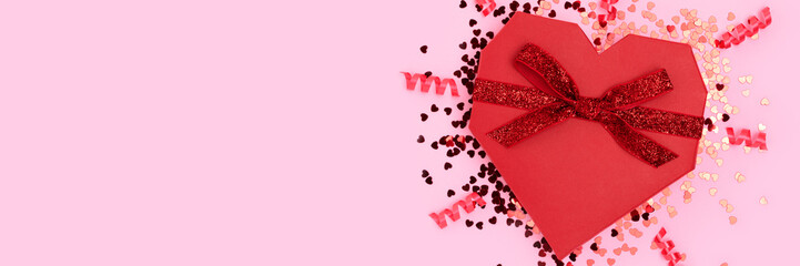 Banner with red gift box in a heart shape tied with glittering ribbon on a pink background.