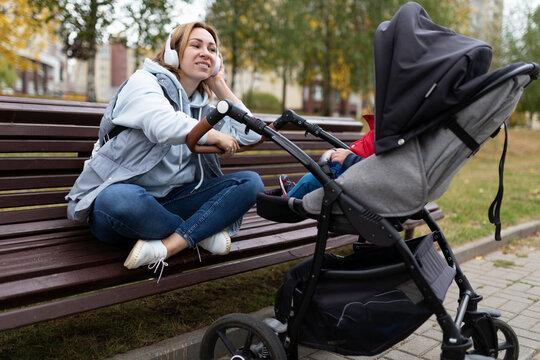 a young mother sits on a bench while walking with a stroller and listens to music on headphones