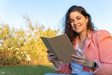 Young woman sitting in a park, holding a book looking and smiling at the camera.