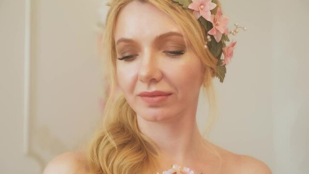 Close-up portrait happy adult blonde girl, holds in hand small pink bouquet flowers inhales smell scent aroma, strokes perfect skin lips body, floral dress. Joyful woman smiling face. wreath hairpin