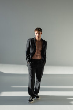 full length of shirtless man in black suit and sneakers standing with hands in pockets on grey background with lighting.