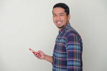 Side view of adult Asian man smiling at the camera while holding his mobile phone