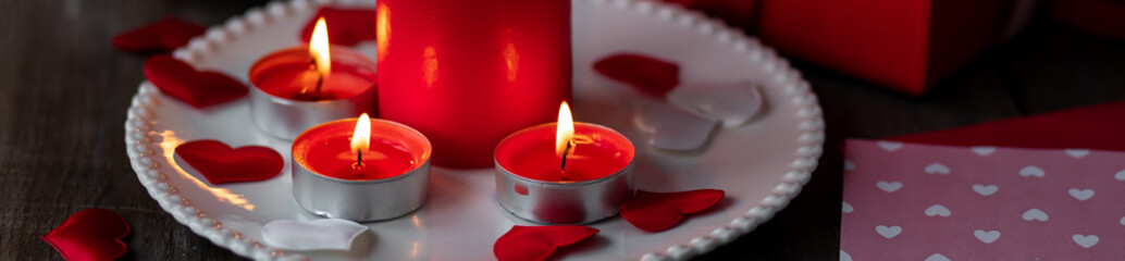Saint Valentine's Day celebration. Red burning candles, hearts, gift box, postcard on dark wooden background. Happy holiday. Table decor for festive dinner, romantic atmosphere. Banner