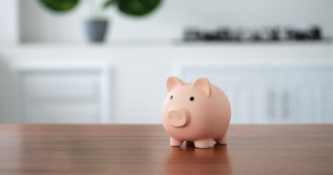 on the wooden table in kitchen is a piggy bank in a bright space. A woman's hand puts a coin in an peachy open piggy bank. The concept of saving money in a piggy bank or new start up. straight view.