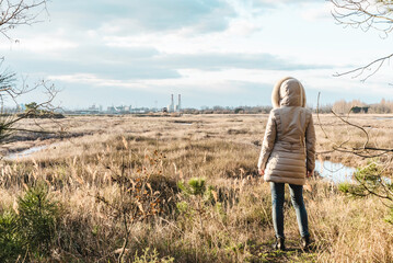 Back view of woman wearing winter clothes in front of grassland wilderness and factories on the...