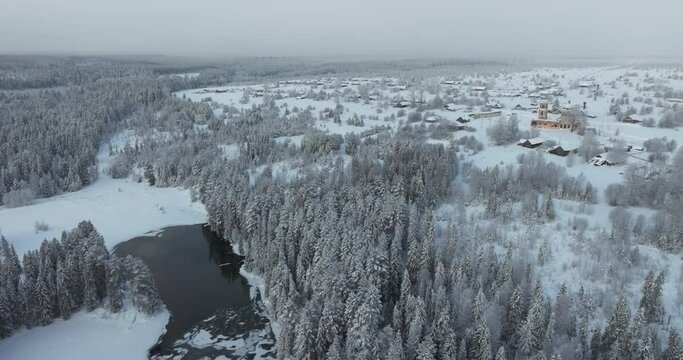Northern winter, the village on the edge of a cinematic dense forest, the spruces and the river covered with a layer of snow. Winter Xmas muted tones drone footage