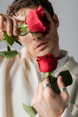 stylish man in white jacket and silver finger rings obscuring face with red roses isolated on grey.