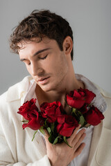 brunette man in white soft jacket standing with closed eyes and holding bouquet of red roses isolated on grey.