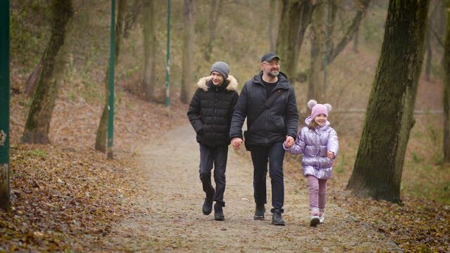 Front view of adult father and little fanny daughter with teenager son walking along autumn forest path cloudy day holding hands and talking. Concept of joint family pastime, parenting and upbringing