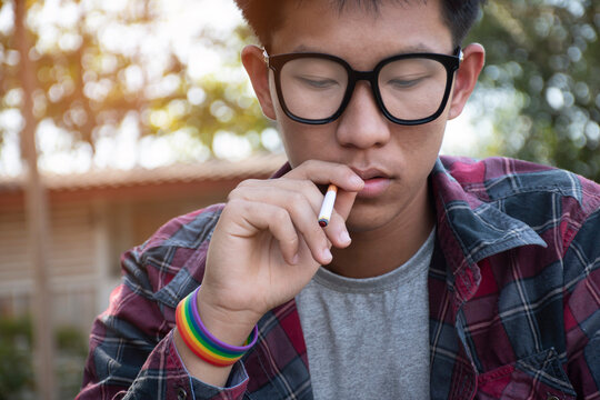 Young Asian Teen Boy In Plaid Shirt Wears Rainbow Wristband And Holds Cigarette In Hand And Smoking, Blurred Background, Concept For Bad Behavior Of Young Teens All Over The World.