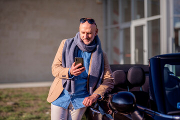 Casual mature man using mobile phone and standing next to his convertible car