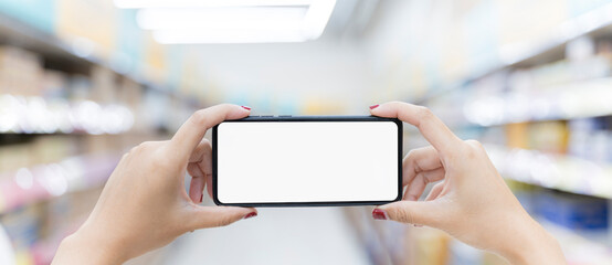 Horizontal close-up shot of female hand holding smartphone, blank screen, background as many shelves.