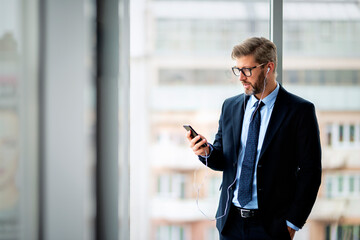 Middle aged businessman using earphone and mobile phone while standing in a modern office