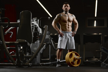 Fototapeta na wymiar Portrait of young muscular man posing shirtless in gym indoors with barbell equipment. Relief body shape. Concept of sport, workout, strength