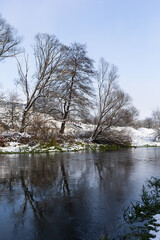 The river flows through the forest. In late fall in the forest the trees stand leafless and reflected in the water of the river After the snowfall, snow lies on the river banks and on the trees