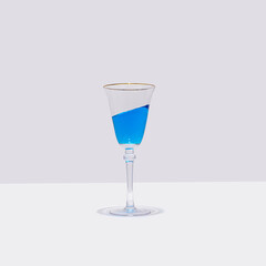 A glass of blue cocktail stands straight with the drink  in it is tilted at an angle. Surreal drinking culture, summer party or celebration composition.