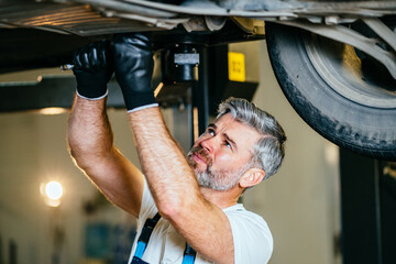 Close up of confident professional handsome mature male mechanic repairing the bottom of the car...