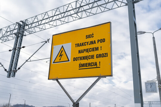 Warning sign with text in Polish language, means overhead electrical contact power lines, danger of death. High voltage hazard. Electric train cables wires, yellow board at railway station platform.