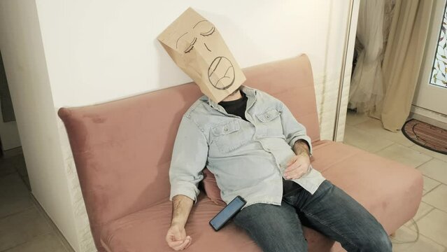 A man wearing a breadbag face, sitting on the sofa at home, falling asleep while browsing on his smartphone.
