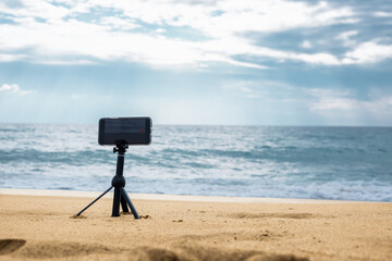  A smartphone camera on a tripod in the foreground stands on the seashore in cloudy weather. High...