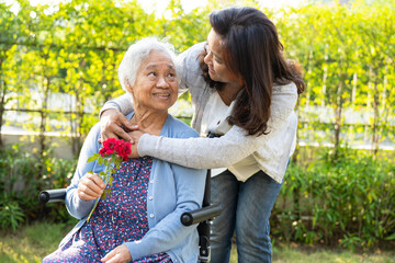 Caregiver daughter hug and help Asian senior or elderly old lady woman holding red rose on wheelchair in park.