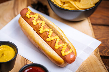 Classic fast food hot dog served with mustard on a rustic wooden board. Served with potato chips...