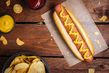 Classic fast food hot dog served with mustard on a rustic wooden board. Served with potato chips and sauces. 