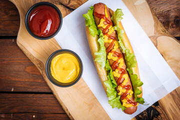 Hot dog with mustard, ketchup and green salad in bun, served with potato chips on a rustic wooden board. Fast food and delicious.