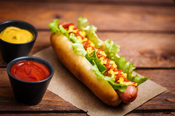Hot dog with mustard, ketchup and green salad in bun, served with potato chips on a rustic wooden board. Fast food and delicious.