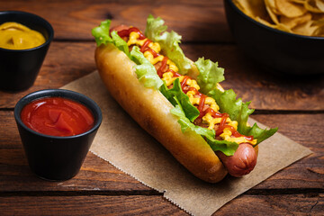 Hot dog with mustard, ketchup and green salad in bun, served with potato chips on a rustic wooden board. Fast food and delicious.