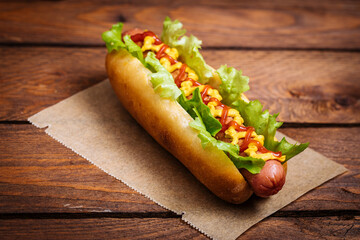 Hot dog with mustard, ketchup and green salad in bun, served with potato chips on a rustic wooden board. Fast food and delicious.