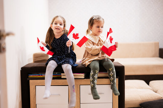 Two Sisters Are Sitting On A Couch At Home With Canadian Flags On Hands. Canada Children Girls With Flag .