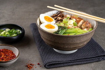 Asian noodle soup, ramen with chicken breast, vegetables, mushrooms and egg in bowl on a slate board on a gray concrete background. Front view from above, healthy eating concept