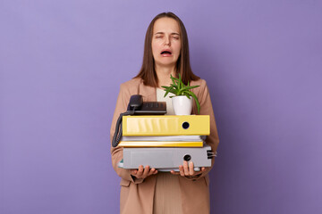 Portrait of depressed stressed Caucasian woman wearing official style jacket holding folders isolated over purple background, female lost her job, crying with despair.