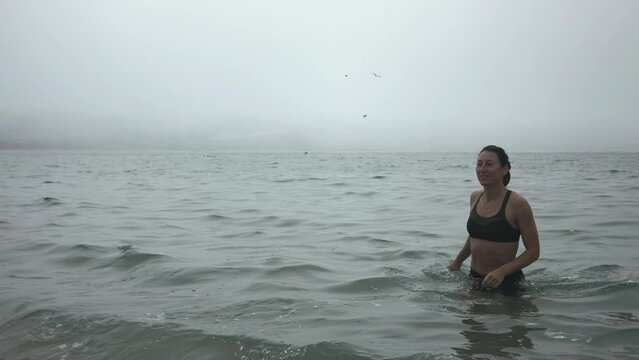 A girl in a black swimsuit confident comes out of the sea into the fog.