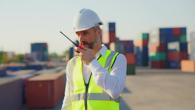 Medium shot of smart handsome Caucasian foreman in white hard hat talking by walkie talkie radio control and communication Industrial with shipping worker. Import and export from abroad.