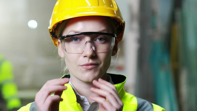 Close up of beautiful Caucasian woman in helmet taking on goggles and smiling to camera. Indoors. Young female worker of plant in hardhat wearing protectional glasses. Protection concept. Portrait.