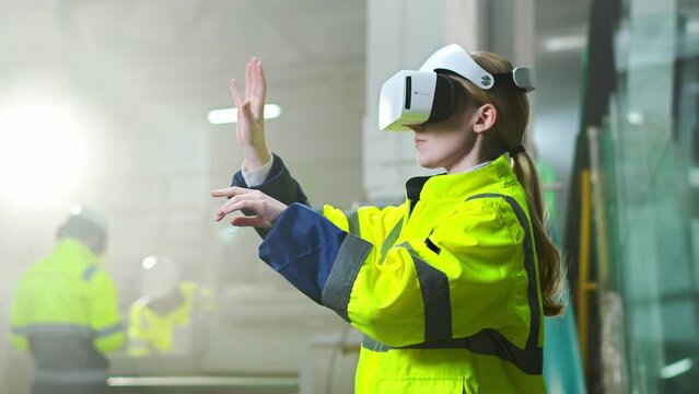 Caucasian female young engineer in headset and yellow uniform standing in factory and having virtual reality experience. Engineering concept. Inside. Blond woman in VR goggles moving hands at plant.