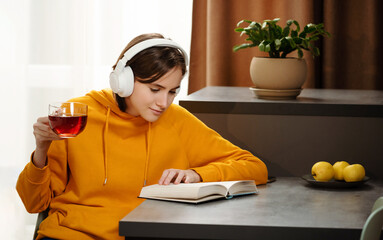 Teen girl sit at the table, reads book and listens music while drinking tea on kitchen. Headphones...