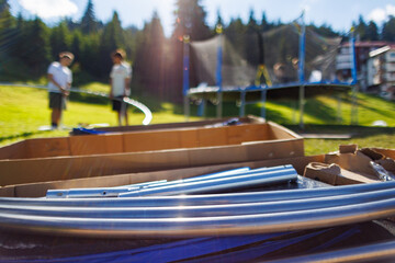Boxes with iron beams to assemble a trampoline as in the background next to people assembling a...