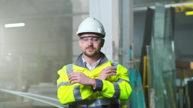 Portrait of handsome Caucasian man in helmet and uniform standing at plant manufactoring and taking on goggles. Indoors. Young worker at factory in hardhat and protectional glasses. Protection.