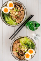 Two bowls of ramen soup with chicken breast, vegetables, mushrooms, pak choi and egg on a white wooden table and chopsticks. Top view, healthy eating concept