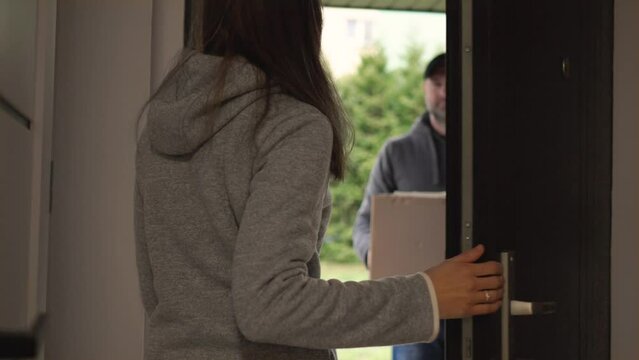 Young woman opens doors of her house and meets delivery man who gives her cardboard box postal package. Concept of courier, home delivery, e-commerce shipping